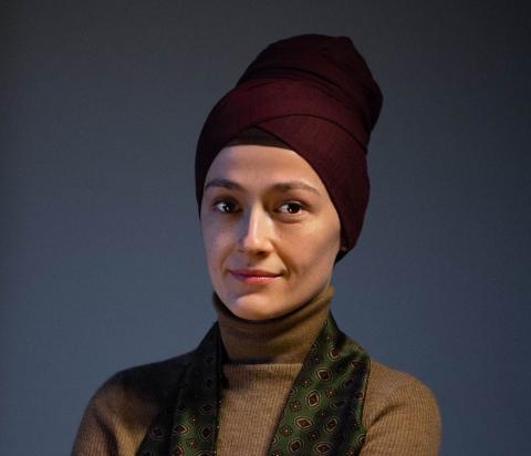 Woman wearing a purple headscarf and beige turtleneck against a dark background